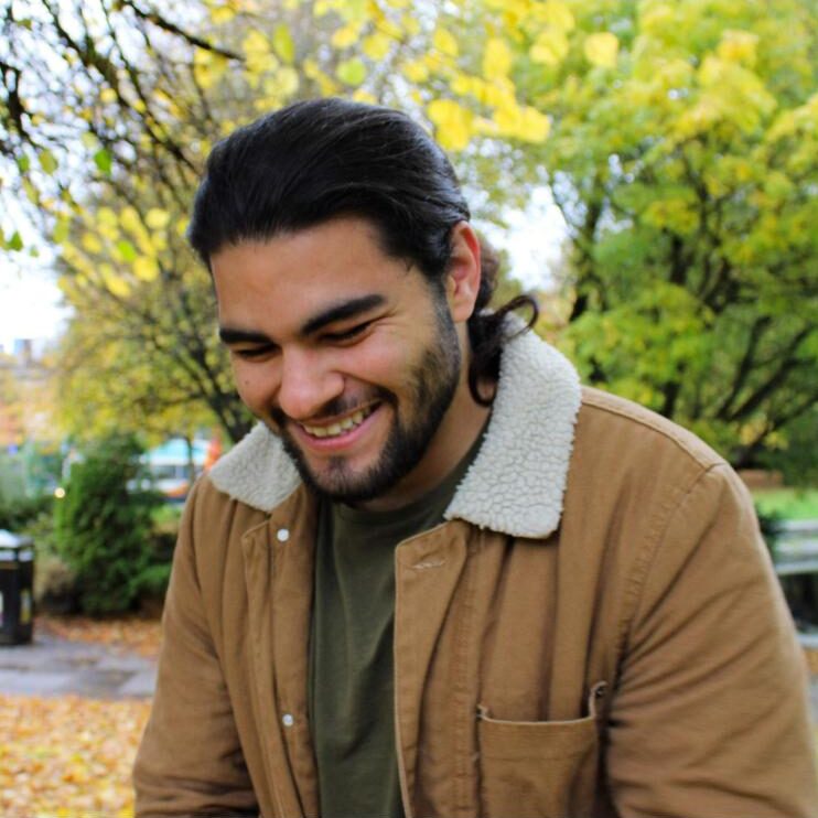A person wearing a brown jacket with a white collar, looking down and smiling in front of some trees.