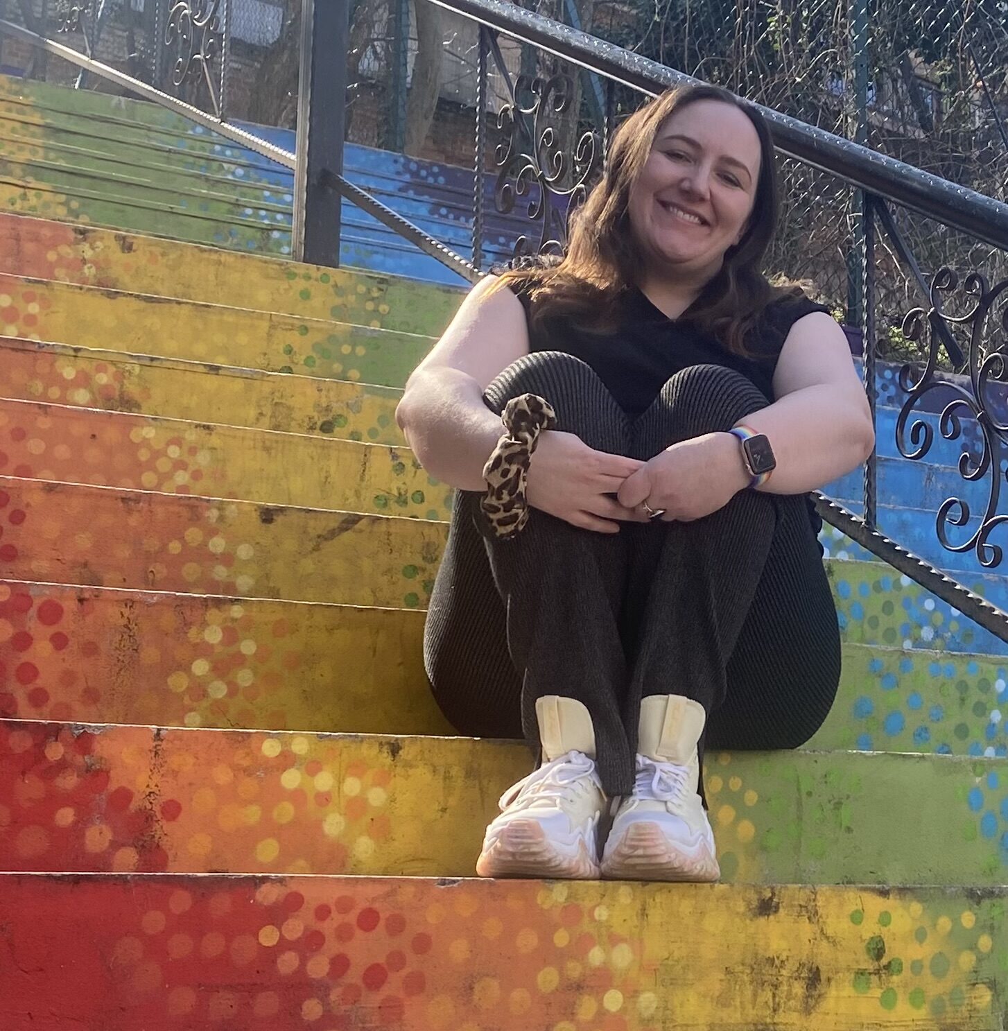 A person sits smiling on brightly painted rainbow-colored stairs with dotted patterns. They are wearing a black top, dark trousers, white shoes, a leopard-print scrunchie, and a smartwatch. Iron railings run along the sides of the stairs, and trees and buildings are visible in the background.